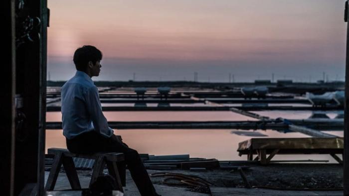 Believer movie: Korean man in white shirt on left looks out over the water while sitting on bench at a dock at sunset