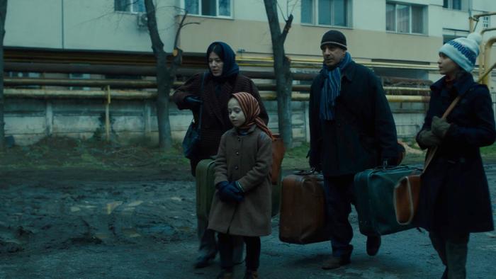 Between Borders: A family of four walks on the street at dusk, holding suitcases 