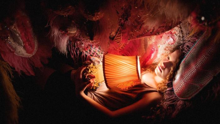 Blaze Movie: Blaze (Julia Savage), holding a small table lamp, curls up in the lap of her imaginary puppet dragon, Zephy