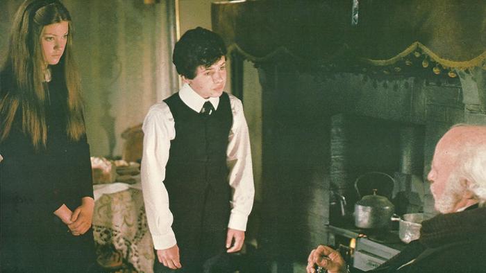The Amazing Mr. Blunden: A young White boy and girl stand in a living space looking down at an old White man as he sits by a fire