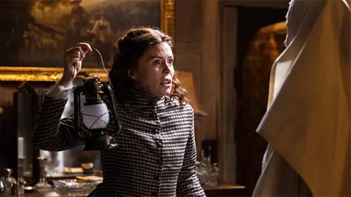 Borley Rectory — The Awakening: An older White woman in 1900 clothing looks frightened as she holds a lantern aloft inside an old manor house