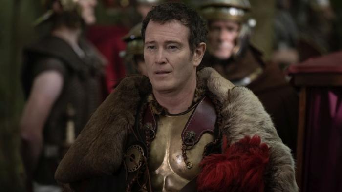Boudica -- Queen of War: A White man with brown hair wears a red Roman commander cloak and gold armor.