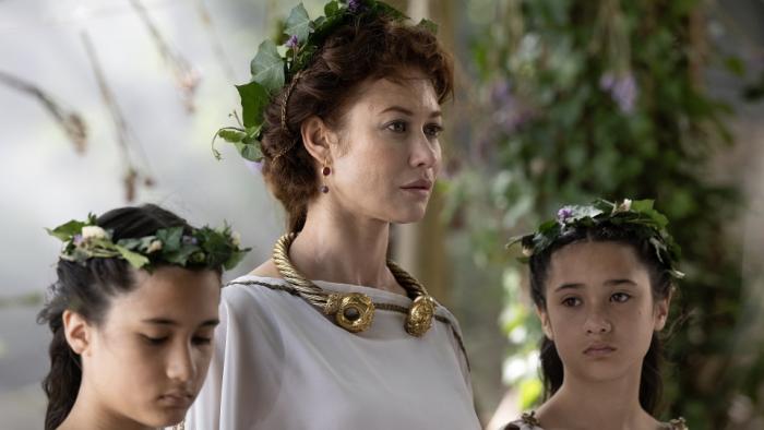 Boudica -- Queen of War: A White brown haired woman in a white robe stands between her two teenage daughters with laurels on their heads.