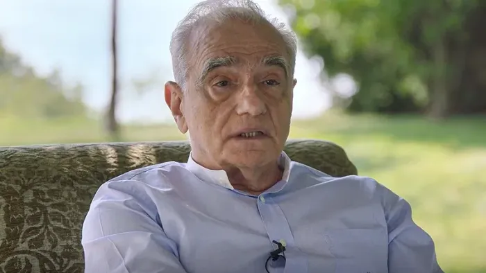 Breakdown: 1975 movie: Martin Scorsese sits on patterned green chair while speaking to the camera, green grass and trees behind him