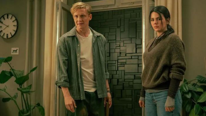 Brick (2025) movie: White German man left center stands next to Costa Rican German woman right, while in their living room