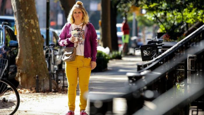 Brittany Runs a Marathon Movie: Brittany stands next to a set of New York City stairs, looking despondent in yellow pants and a purple sweater. 