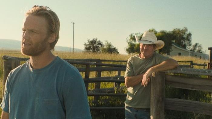 Broke movie: Wyatt Russell in blue t-shirt left talks with Dennis Quaid in white cowboy hat behind him while on their farm, next to a horse paddock