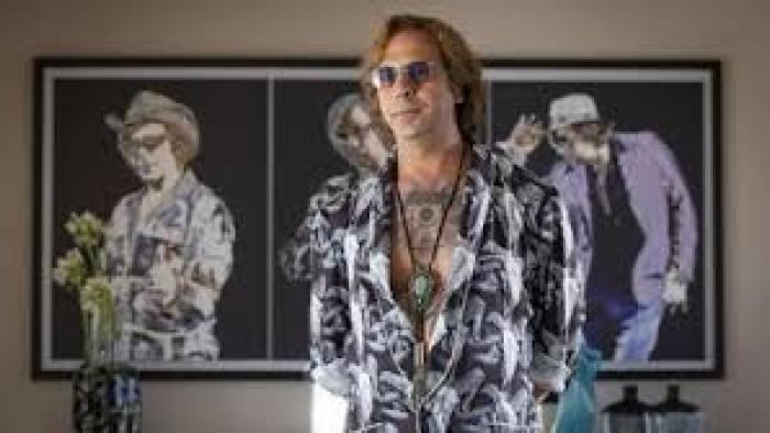 Buen Camino Movie: Man in patterned shirt and sunglasses stands in front of artistic wall portraits.