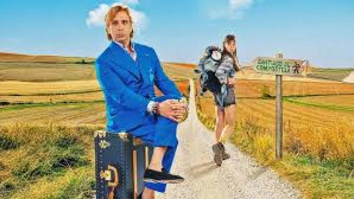 Buen Camino Movie: Man in blue suit sits on suitcase, woman with backpack stands on rural path.