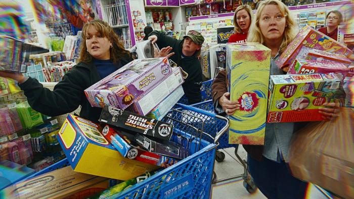 Buy Now! The Shopping Conspiracy poster: Frenzied shoppers push full carts and carry armfuls of items in a busy store