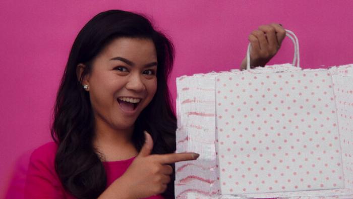 Buy Now! The Shopping Conspiracy poster: A woman smiles and points to a shopping bag against a pink backdrop
