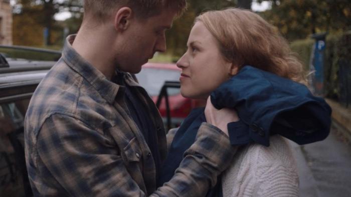 Calibre: A White man holding is pregnant partner with a car in the background
