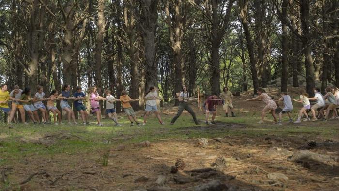 Camp Crasher movie: In woodsy area, preteens play tug-of-war, holding onto a long rope either side of a Spanish woman in middle
