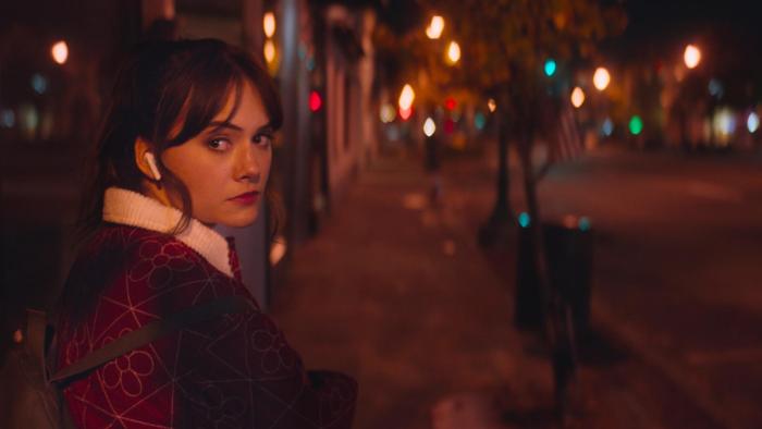 Cat Person Movie: Emilia Jones walks down a desolate street at night, wearing headphones and looking over her shoulder