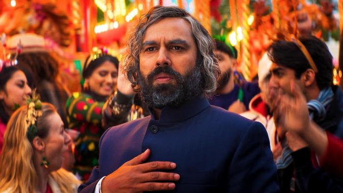 Christmas Karma: Man in dark formal attire at a festive gathering with colorful decorations and crowd in background