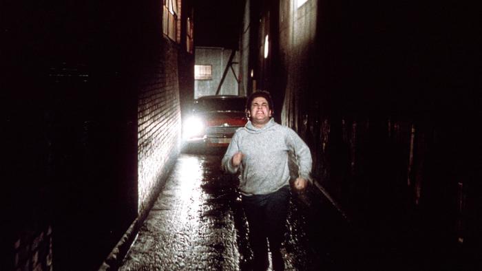 Christine Movie: A man in a gray hoodie runs toward the camera in an alley, sinister headlights looming behind him