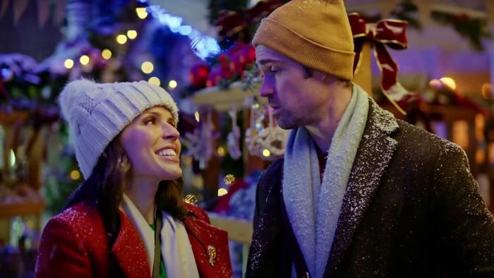 Christmas Plus One: A White woman smiles up at a White man, both wearing coats, scarves, and hats at a Christmas market.
