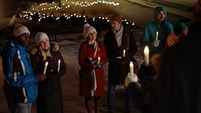 Christmas Plus One: A group of people gather together in coats, hats, and scarves, holding candles with Christmas lights behind them.