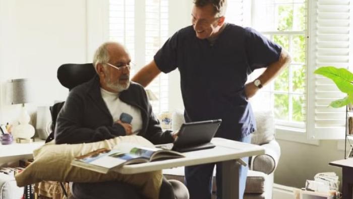 Chronic movie: A smiling White male care-home worker and an older male patient watch a tablet