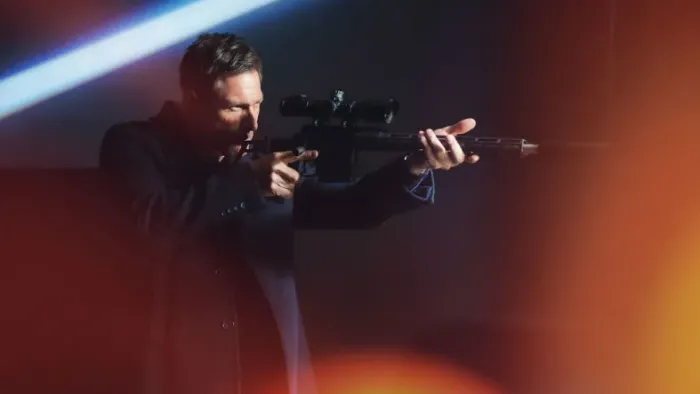 Classified movie: Aaron Eckhart in black leather jacket center walks to the right holding up rifle and looking down the sight