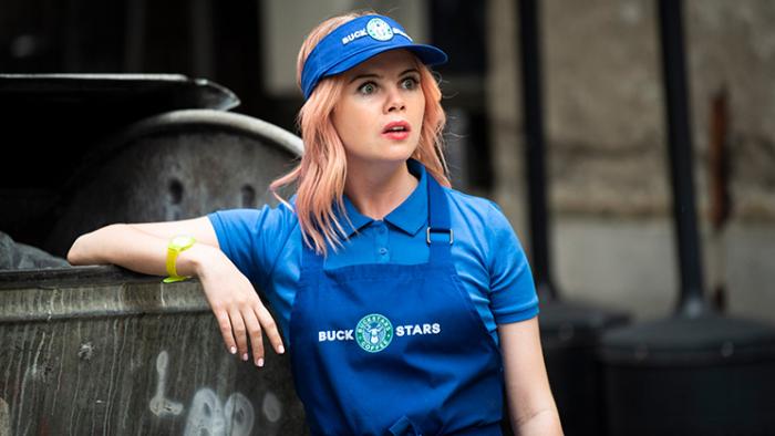 Coffee Wars: A White woman with pink hair in a blue coffee shop uniform leans against a bin