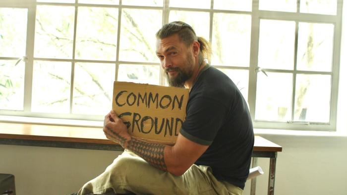 Common Ground Movie: Jason Momoa sits in a classroom holding a cardboard sign with the words "Common Ground" on it