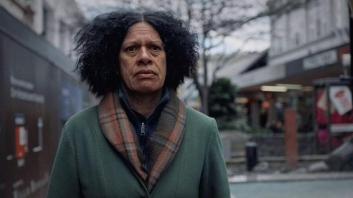 Cousins Movie: An adult Māori woman gazes unhappily from a retail street, wearing a scarf and coat