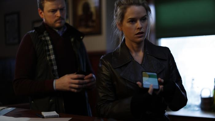 Cult Killer Movie: Private eye Cassie Holt (Alice Eve, right) holds a cell phone and appears to be questioning someone off-camera, while officer Rory McMahon (Paul Reid) stands behind her