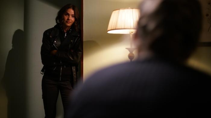 Cult Killer Movie: Revenge-driven Jamie Douglas (Shelley Hennig) leans against a wall in Cassie's apartment, speaking to Cassie, who we can see out of focus and from the back