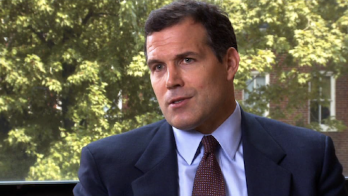 Inside Job: A White man wears a suit and sits in front of a leafy view from a window as he's interviewed