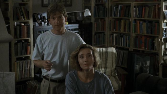 Dead Again: Emma Thompson sits in a chair in a dimly lit living room and Kenneth Branagh stands behind her holding a business card
