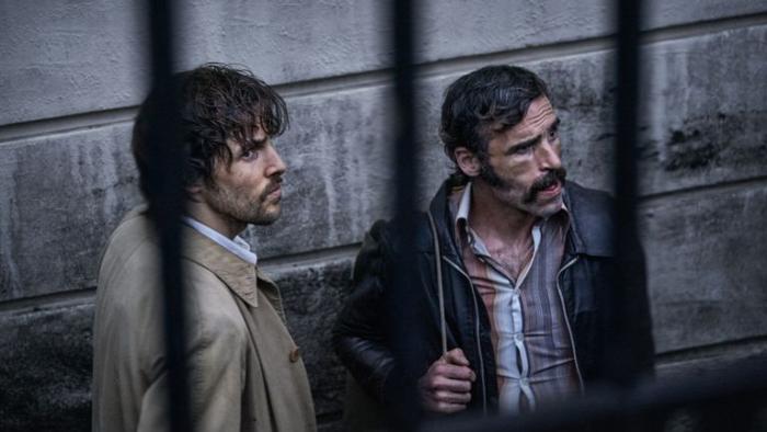 Dead Shot: Two White men are seen through bars waiting next to a building