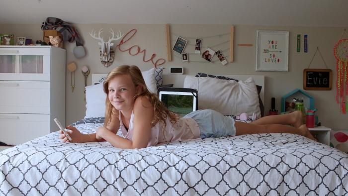 Dear Thirteen movie: Evie lying on bed, smiles and holds a smartphone, wearing white sleeveless top and blue shorts