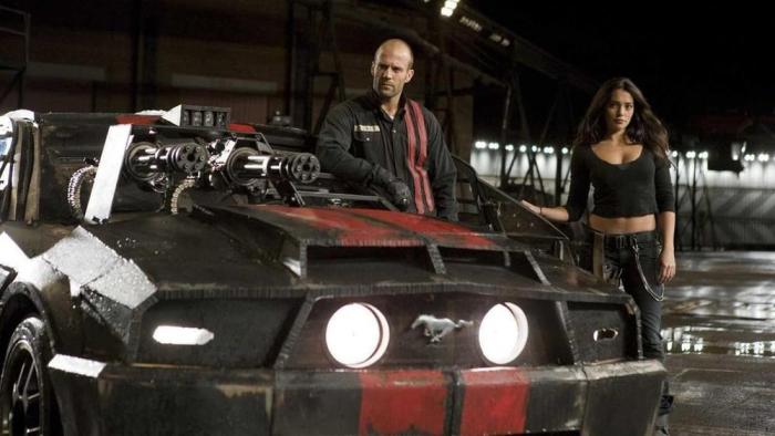 Jason Statham and Natalie Martinez stand behind a black and red sports car.