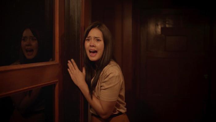 Death Whisperer 2 movie: Indonesian woman with long black hair center inches along hallway wall yelling, her reflection on left in glass window
