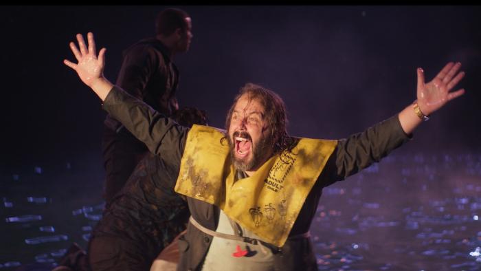 Deep Water Movie: Wearing a yellow life vest in a life raft, Dan (Angus Sampson) signals by raising his arms and shouting