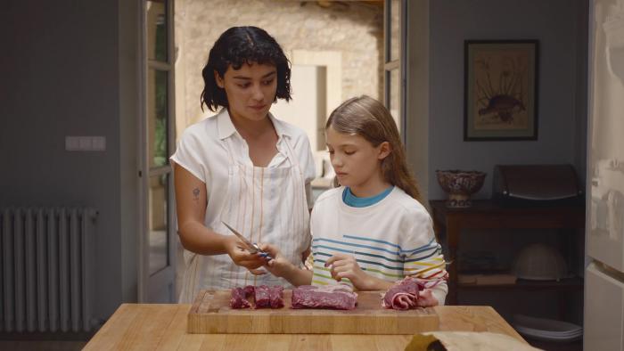 Delicious movie: Spanish woman in white shirt and apron hands boning knife to blond girl while they stand over kitchen island with meat on it