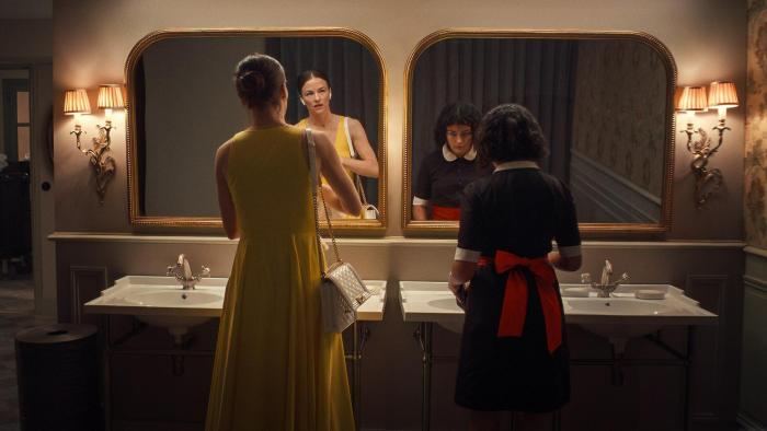 Delicious movie: German white woman in yellow dress left looks in mirror while in fancy bathroom, a Spanish woman in black maid outfit to her right