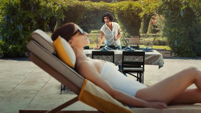 Delicious movie: White German woman in white bathing suit lies back on poolside chair while Spanish woman sets patio dining table in background