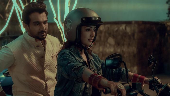 Dhoom Dhaam movie: Indian man in white suit gets on back of motorcycle driven by Indian woman in jean jacket, red bangles, and helmet center