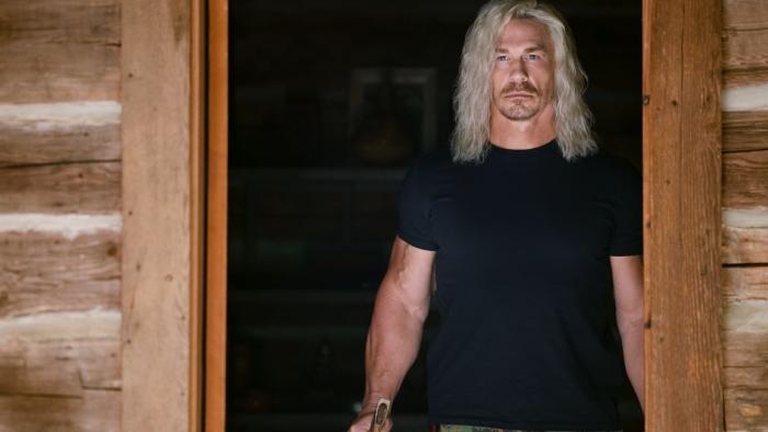 Die Hart 2: Die Harter movie: John Cena in black t-shirt and long blond hair stands under wooden cabin doorway