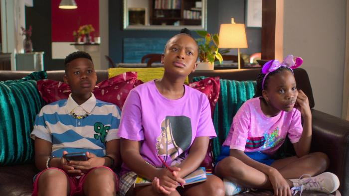 Disaster Holiday movie: Three South African Black children (boy left, girls center and right) wearing colorful clothing sit on couch in living room