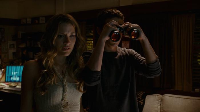Sarah Roemer and Shia LaBeouf stand in a dark room. Shia is holding a pair of binoculars up to his face.