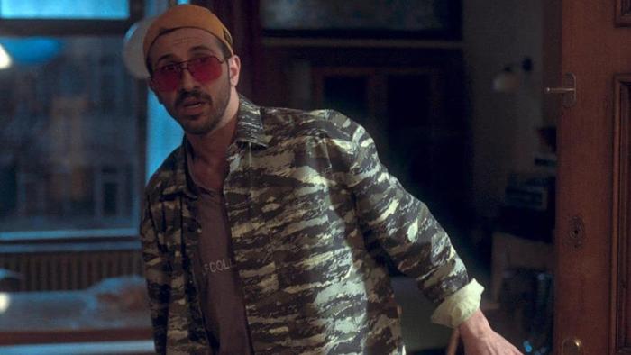 Don't Leave movie: white man center wearing orange beanie, red sunglasses, and camo jacket looks worried