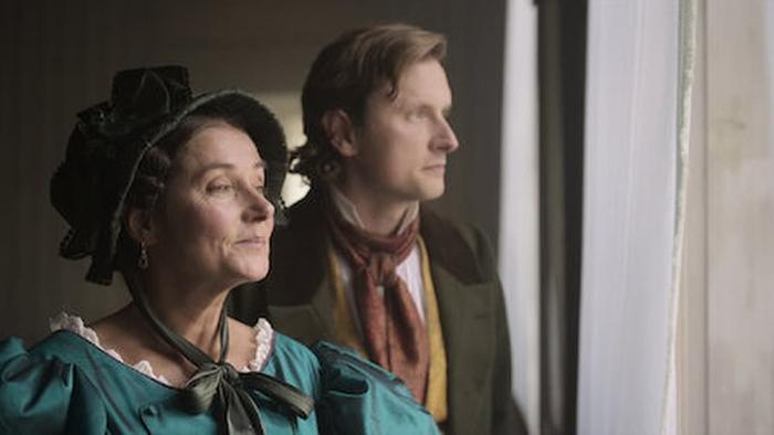 Ehrengard: The Art of Seduction movie: A Danish white woman in teal dress and black bonnet left and a Danish white man in red scarf and suit jacket center look out the window