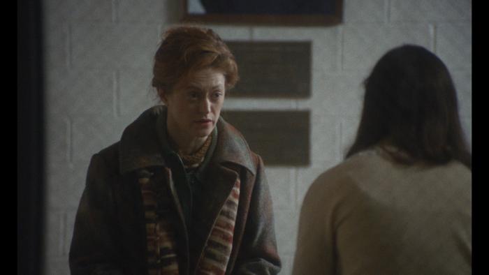 Eileen Movie: A middle-aged White woman speaks to a young woman in a grey, bleak building