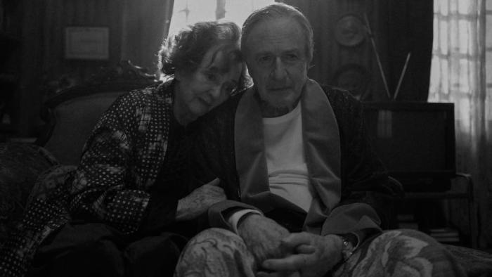 El Conde movie: A Chilean older woman in polkadot pajamas left sits on a couch next to a Chilean older man in robe and white shirt right with windows and an old television behind them