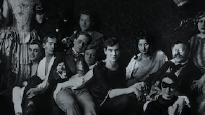 Still from Eldorado: Everything the Nazis Hate: Bespectacled Magnus Hirschfeld (far right), founder of  the world's first gay rights organization in 1897, Institute for Sexual Science in Berlin, Germany and queer individuals gather together wearing attire that includes gowns, suits, and facial masks