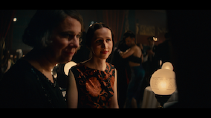 Still from Eldorado: Everything the Nazis Hate: Queer couple Toni Ebel (left) and Charlotte Charlaque seated at a nightclub table while in conversation with others at Berlin, Germany's Eldorado, a hub for gays, lesbians, and trans individuals during the 1920s, wearing dark-colored tops