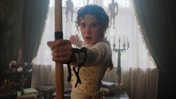Enola Holmes movie: Enola holds up a bow and arrow indoors